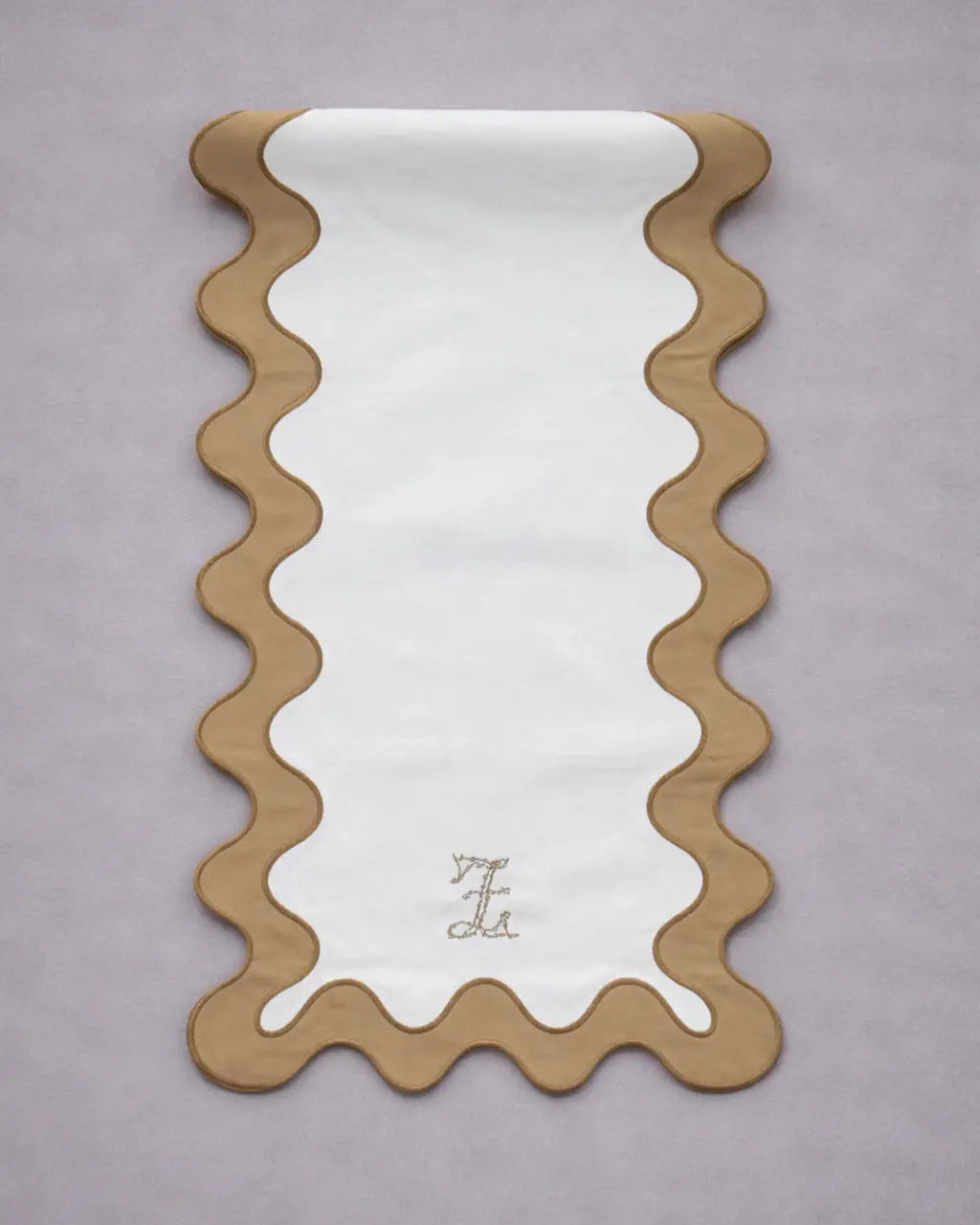 Chemin de table rectangulaire en tissu clair, bordé d’un large liseré ondulé couleur bronze. Le motif forme des vagues régulières tout autour. Au bas du chemin de table, un petit monogramme brodé est visible au centre. L’ensemble est posé sur un fond gris clair.