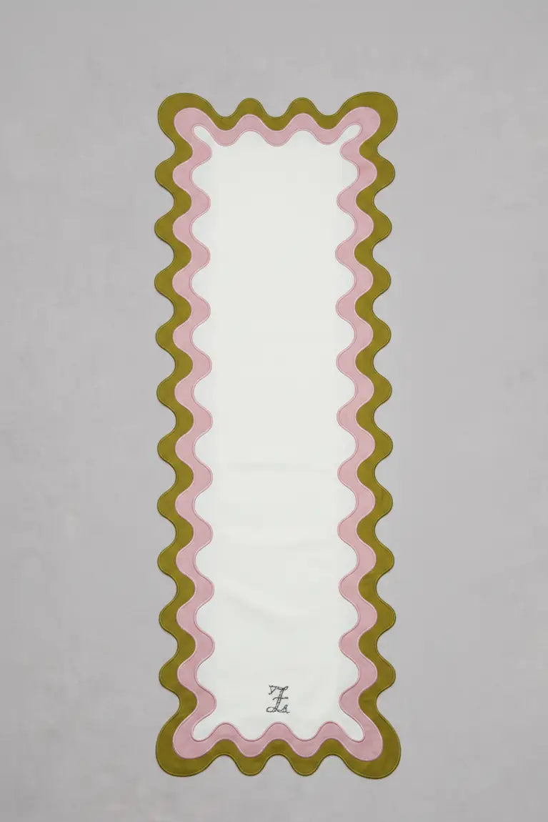 Chemin de table rectangulaire en tissu clair, bordé d’un double liseré ondulé vert olive et rose pâle formant des courbes régulières sur les côtés et en bas. Un monogramme brodé est centré en partie inférieure. Le chemin de table est présenté sur un fond gris clair.
