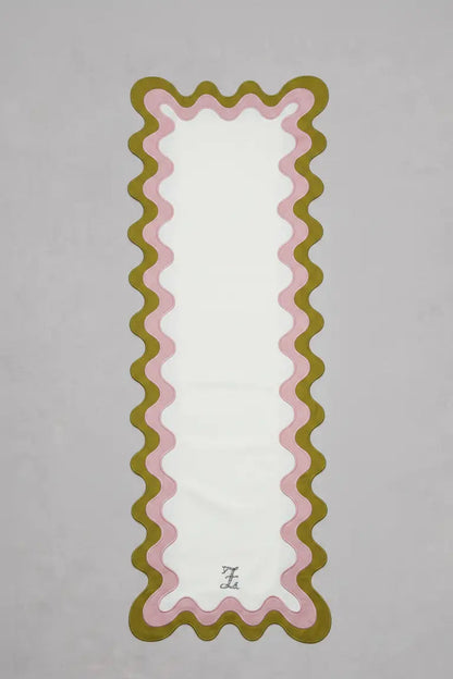 Chemin de table rectangulaire en tissu clair, bordé d’un double liseré ondulé vert olive et rose pâle formant des courbes régulières sur les côtés et en bas. Un monogramme brodé est centré en partie inférieure. Le chemin de table est présenté sur un fond gris clair.