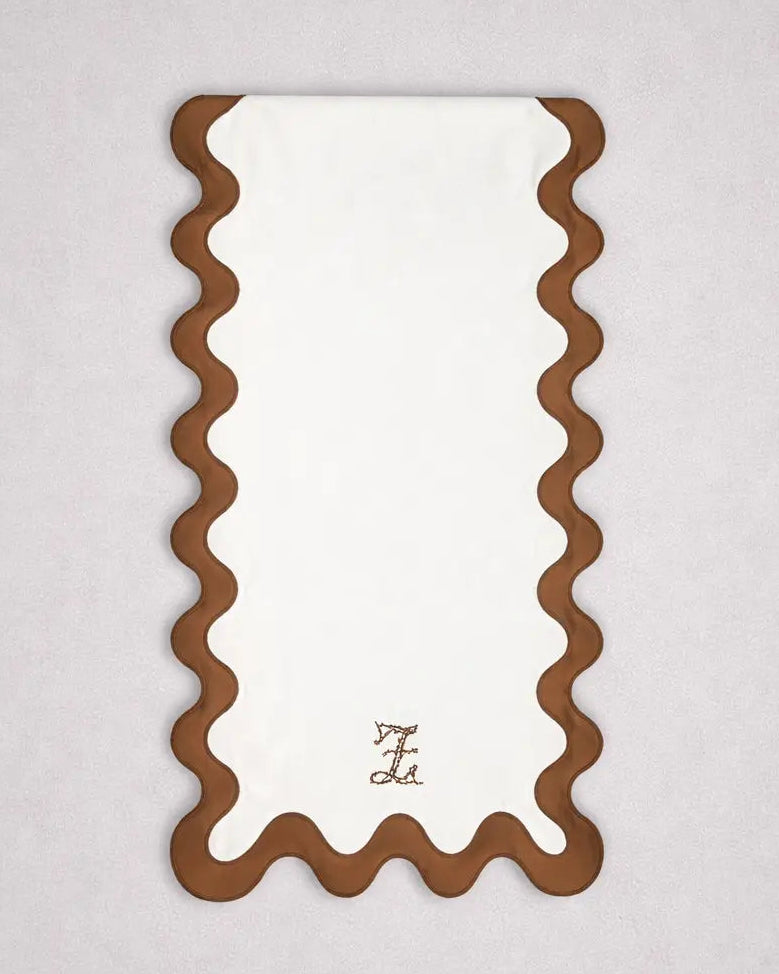 Chemin de table rectangulaire en tissu clair, bordé d’un liseré ondulé marron dessinant des courbes régulières sur les côtés et en bas. En partie inférieure, un monogramme brodé est centré. Le chemin de table est présenté sur un fond gris clair.