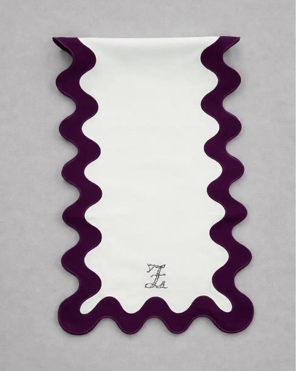 Chemin de table rectangulaire en tissu clair, bordé d’un liseré ondulé couleur prune dessinant des courbes régulières sur les côtés et en bas. En partie inférieure, un monogramme brodé est centré. Le chemin de table est présenté sur un fond gris clair.