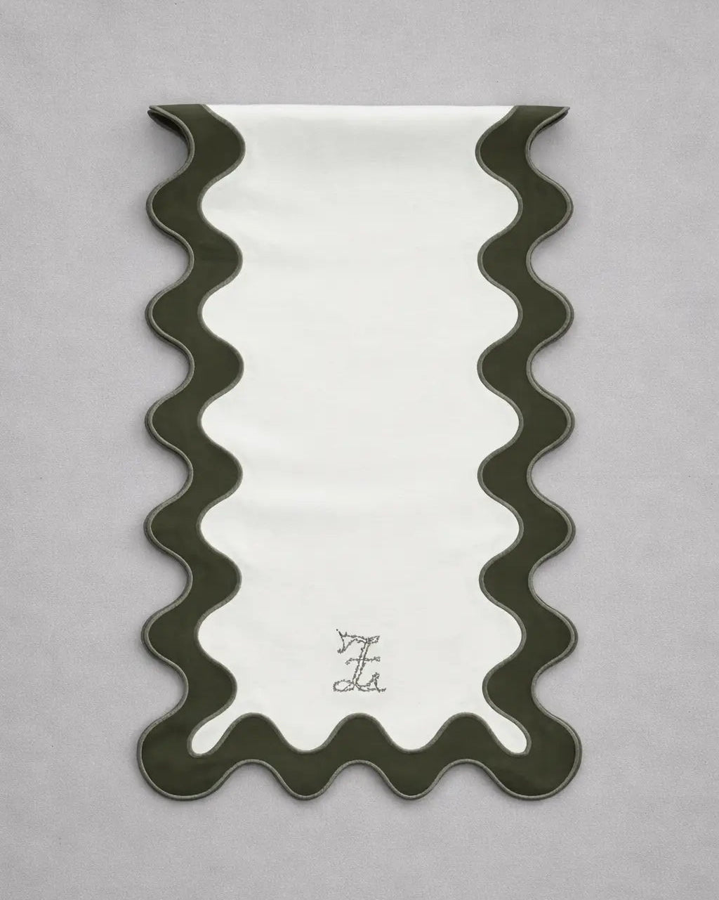Chemin de table rectangulaire en tissu clair, bordé d’un liseré ondulé vert foncé dessinant des courbes régulières sur les côtés et en bas. En partie inférieure, un monogramme brodé est centré. Le chemin de table est présenté sur un fond gris clair.